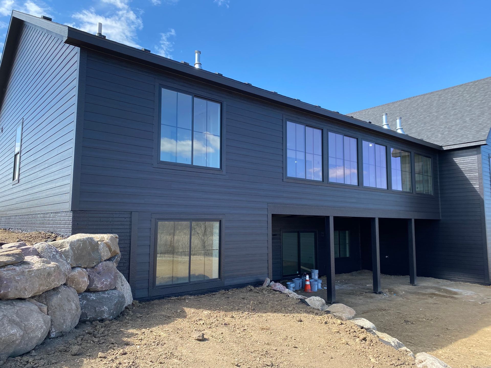 Black modern house exterior with large windows, unfinished yard, and stone wall.
