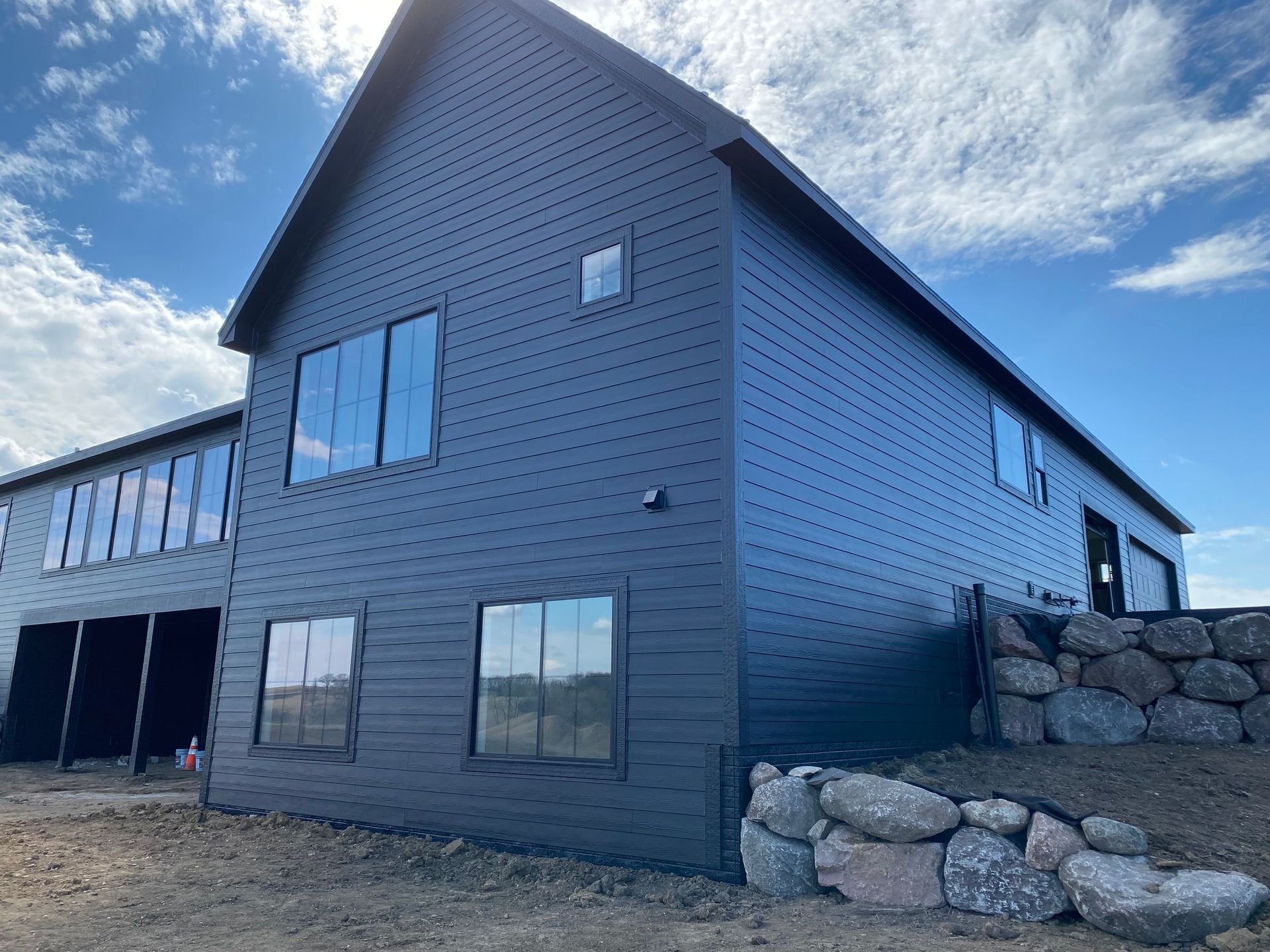 Dark gray modern house with large windows, set on a slightly elevated, rocky landscape. Blue sky overhead.