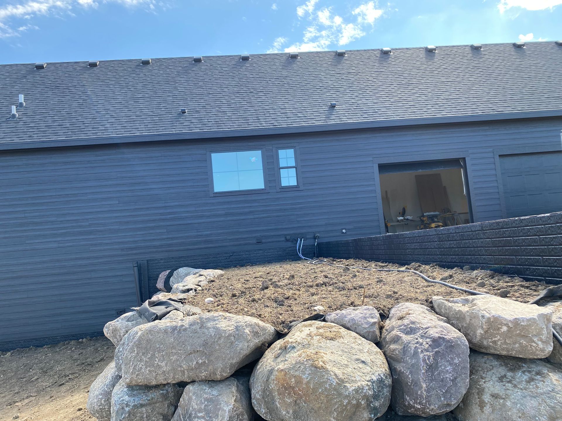 Exterior of a building under construction. Dark siding, gray roof, windows, and large rocks in the foreground.