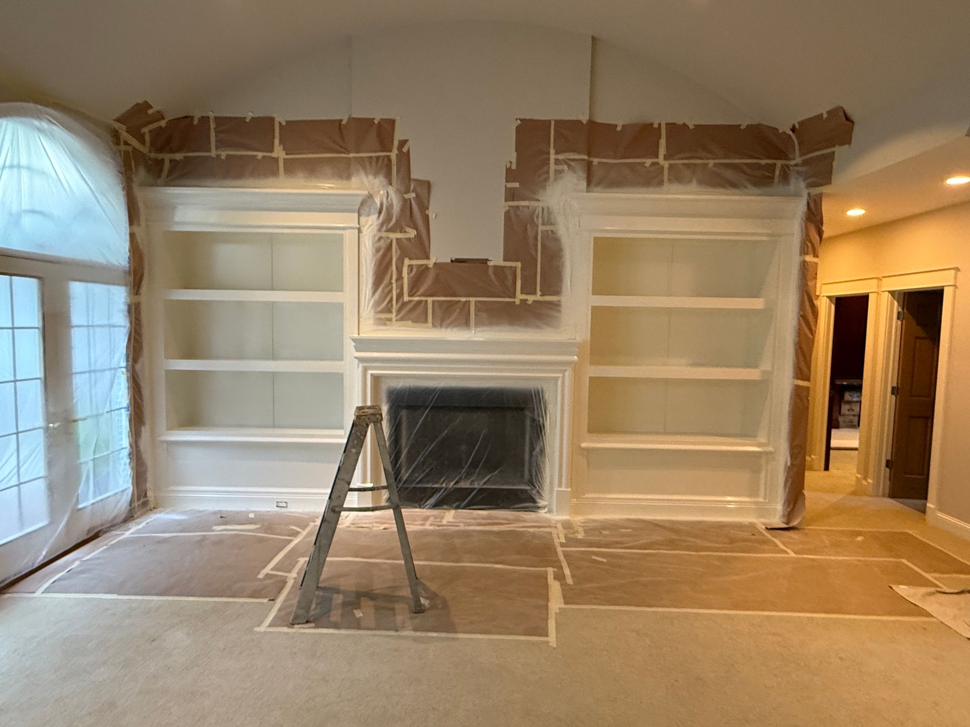 Room with built-in shelves, fireplace, and arched ceiling; being painted. Protective paper and plastic cover surfaces.