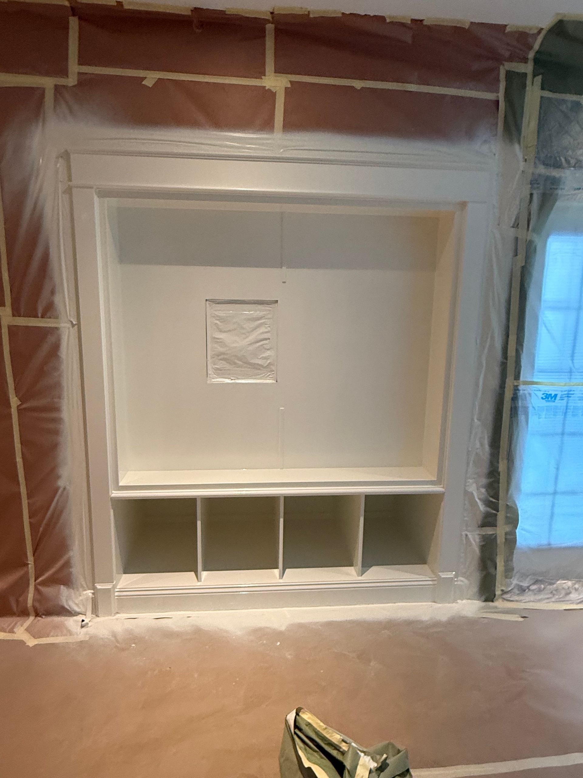 A built-in white cabinet in a wall, with open cubbies and a central indentation. Construction underway, protected by plastic and tape.