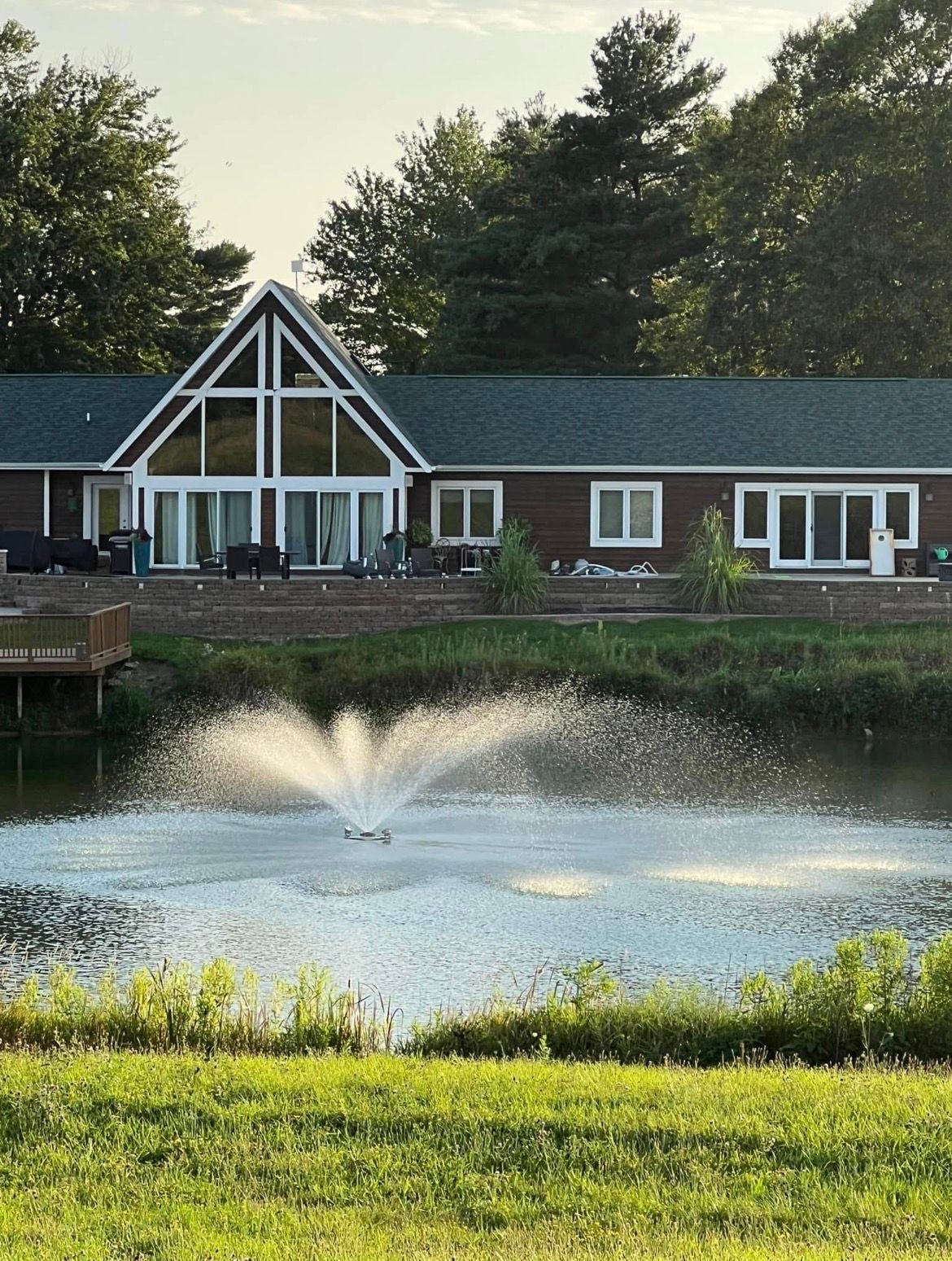 A modern house with a large A-frame window overlooks a pond featuring a spray fountain and a grassy foreground.