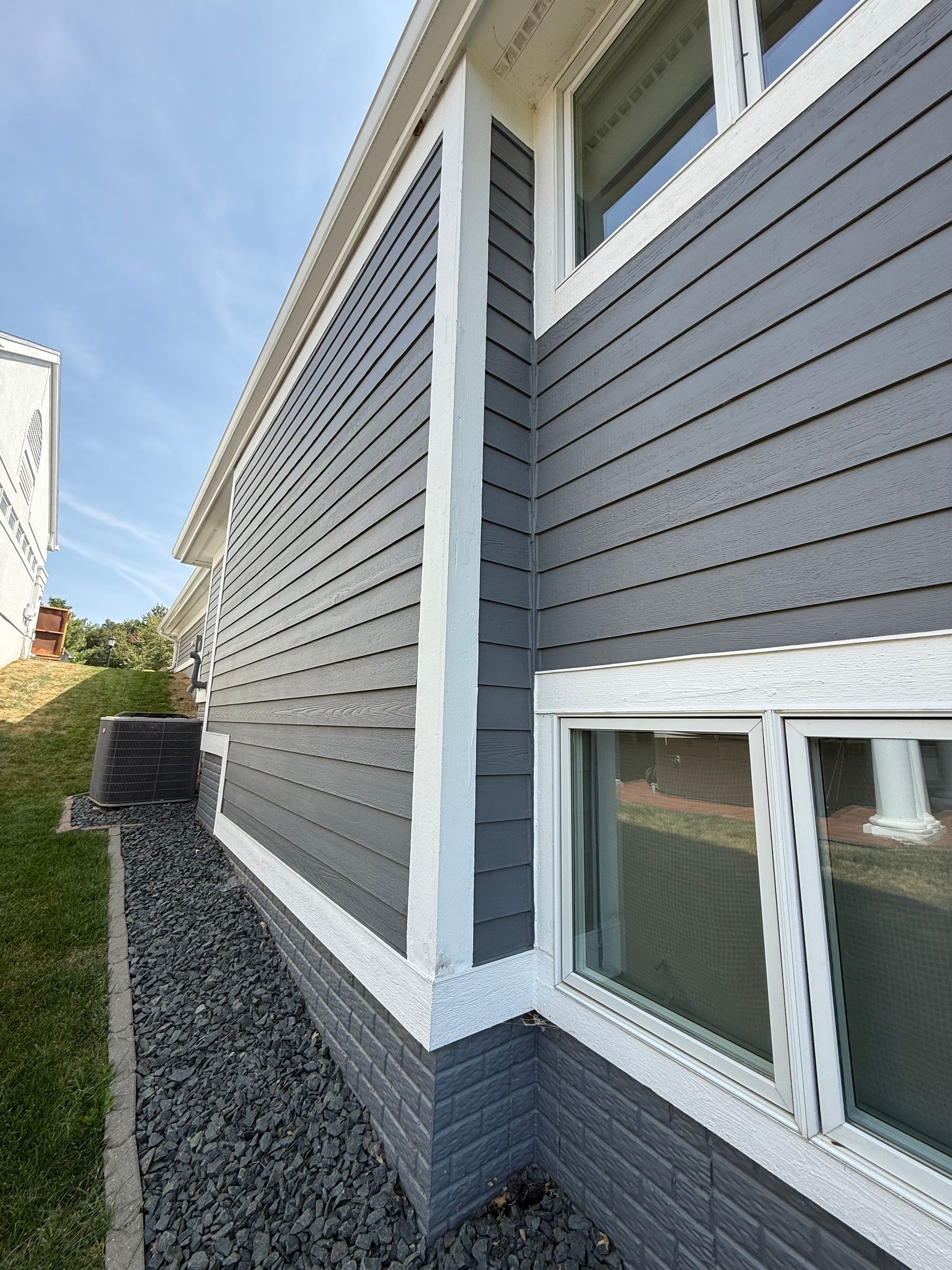Gray siding on a building with white trim and windows, a brick base, and a gravel bed.