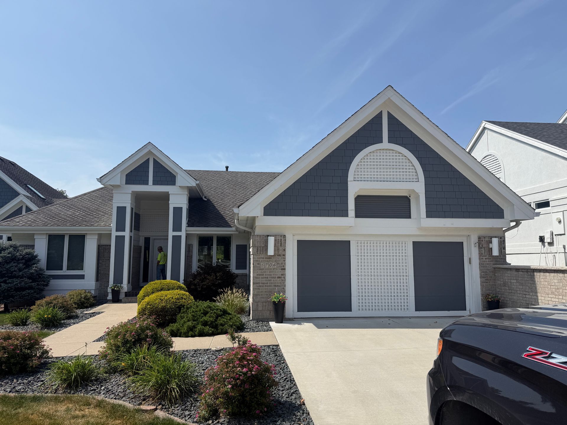 Suburban home with blue siding, gray roof, and stone accents. Driveway with landscaping under a blue sky.