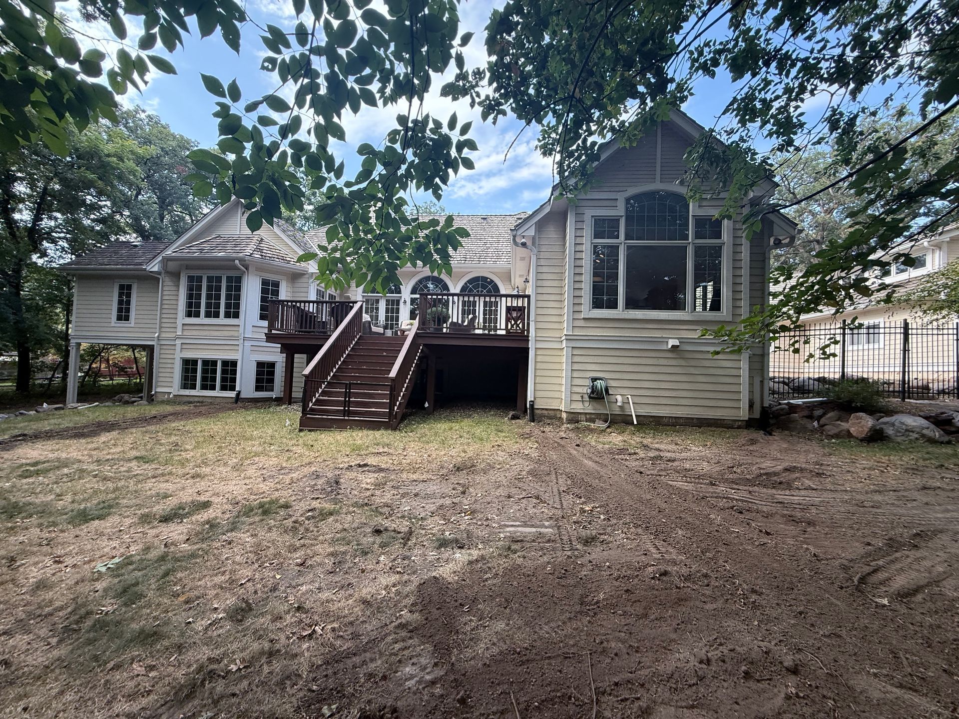 Back of a light-colored house with a dark wooden deck. Overgrown yard, blue sky, trees.