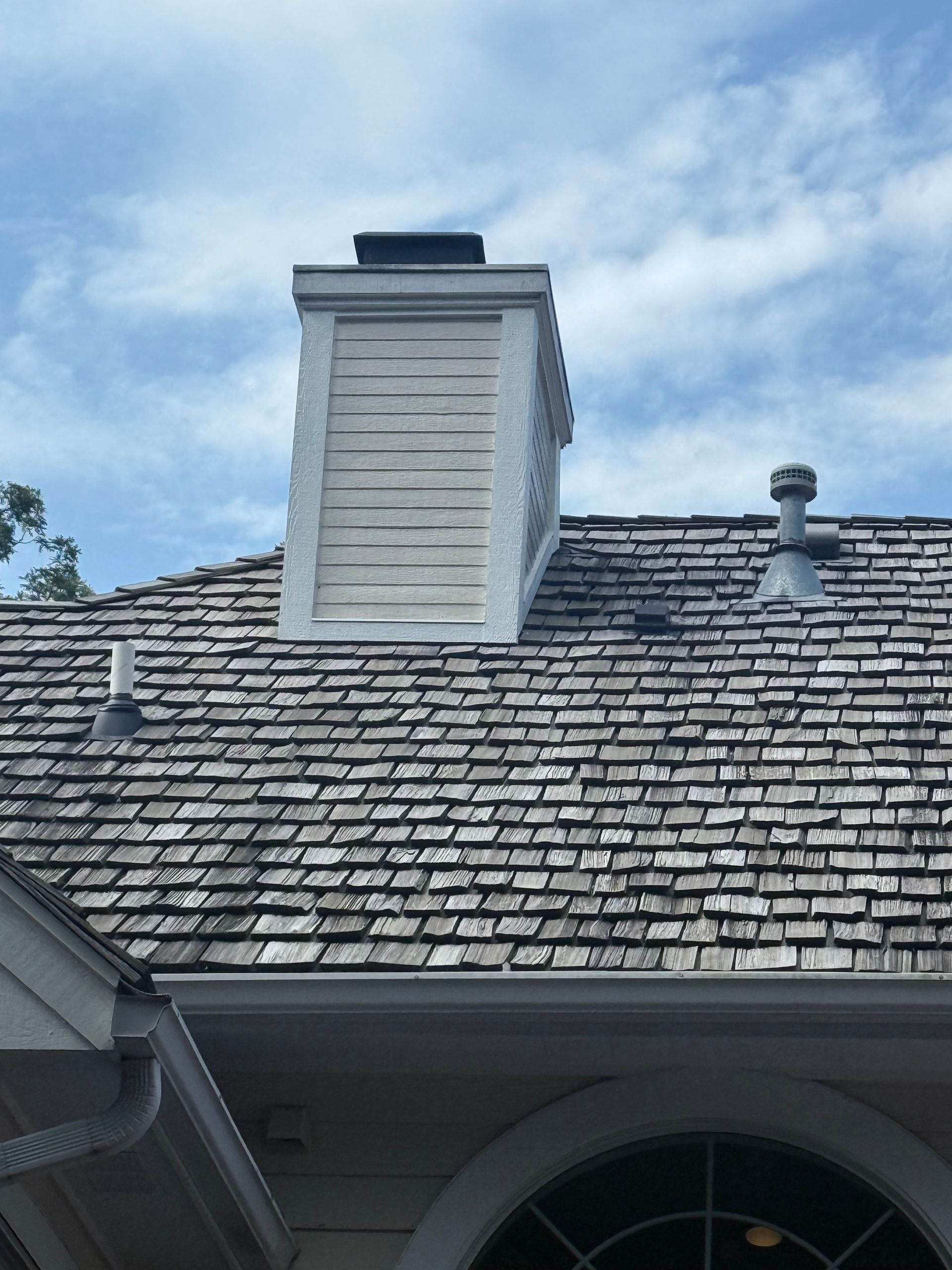 Chimney on a roof with wood shingles against a cloudy blue sky.