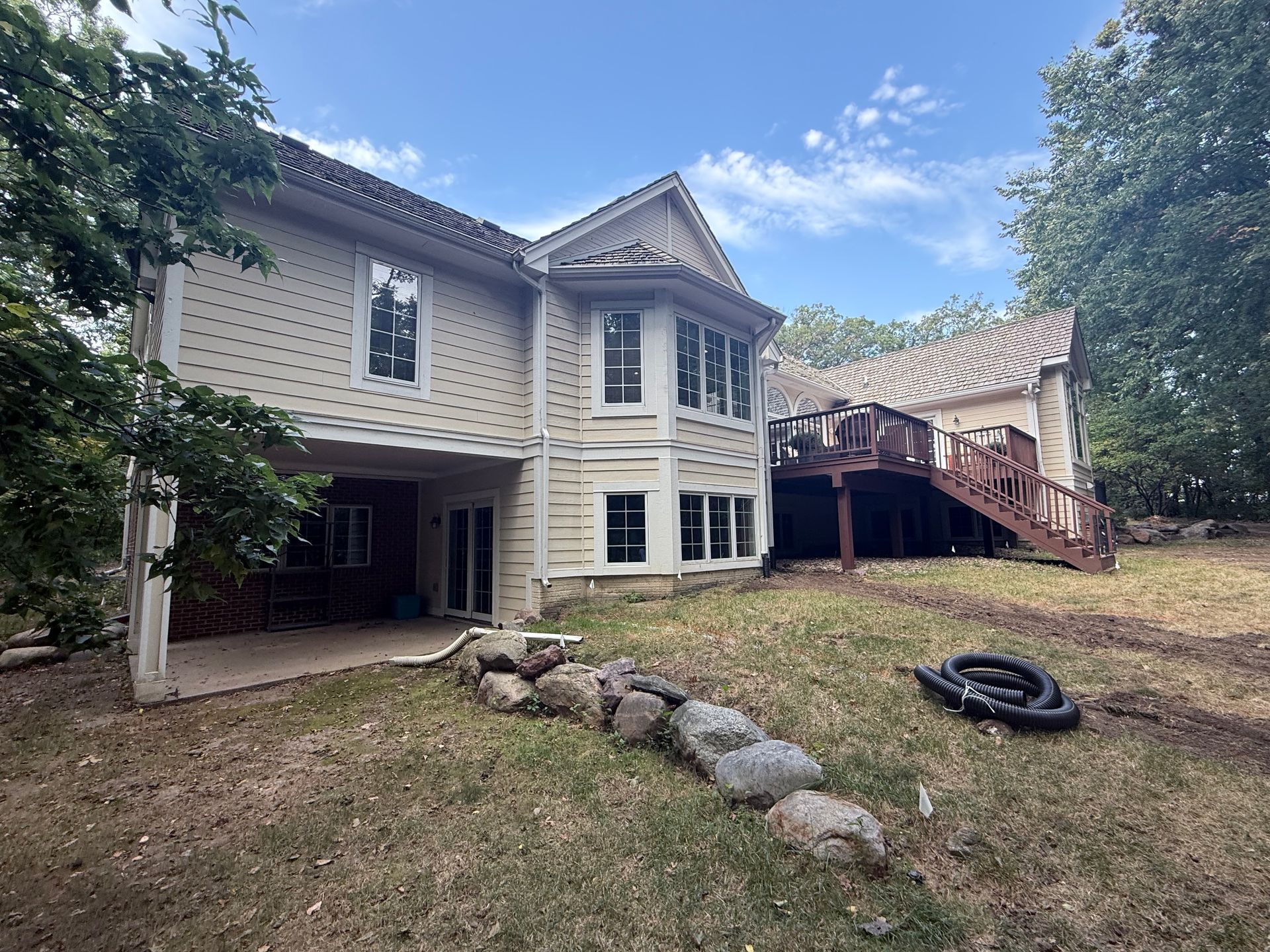 Beige two-story house with a deck and sloping backyard. Blue sky and trees surround the home.