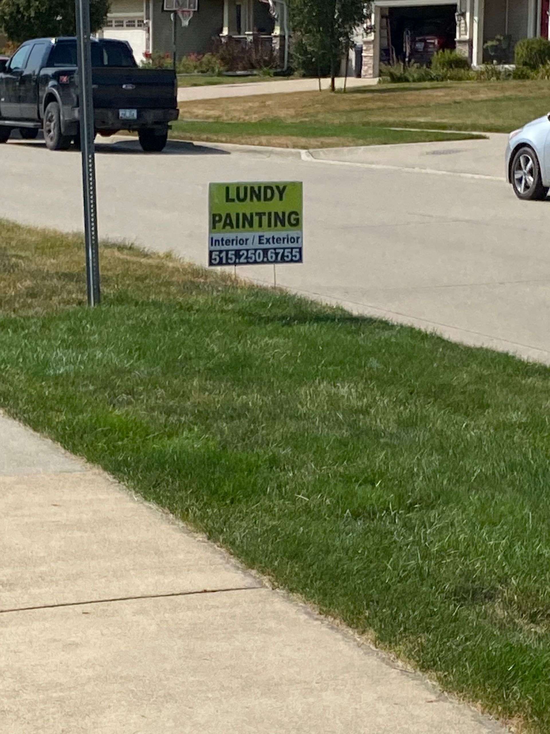 Lundy Painting sign on grassy lawn by sidewalk. Truck and car in background.