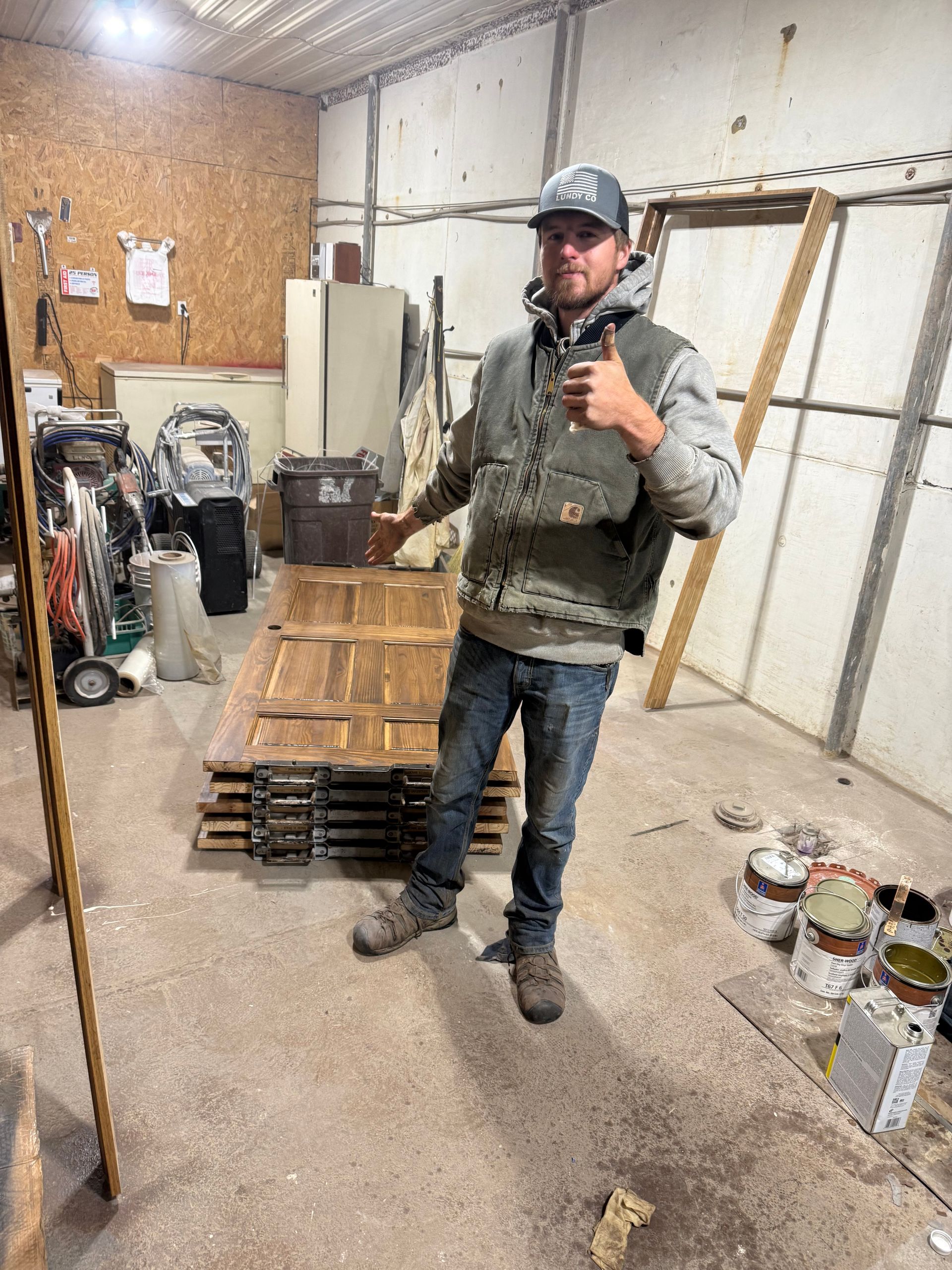 Man in work clothes gives a thumbs up beside a stack of old wooden doors in a garage.