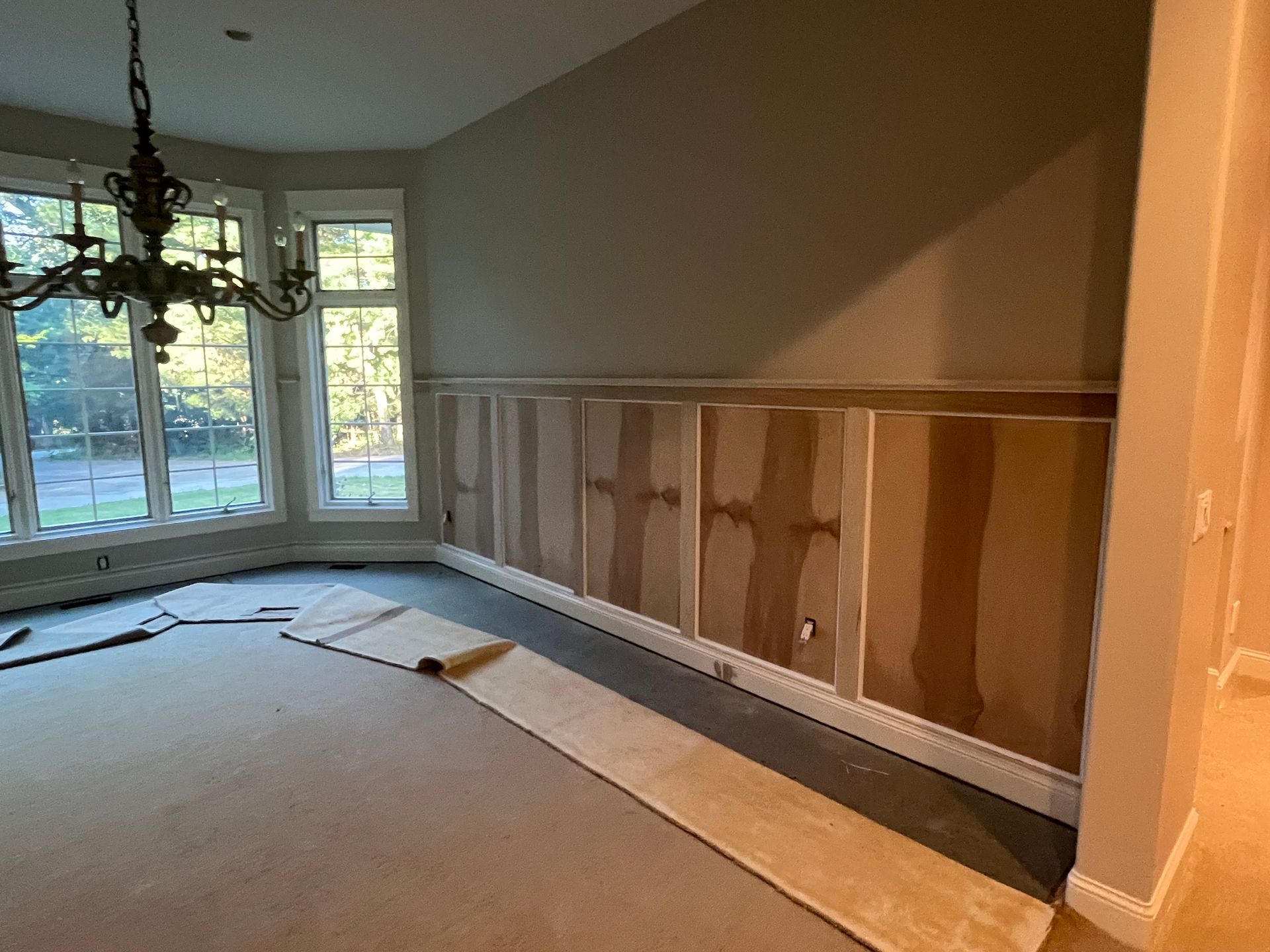 Room interior with wainscoting under construction, carpeted floor, large windows, and a chandelier.