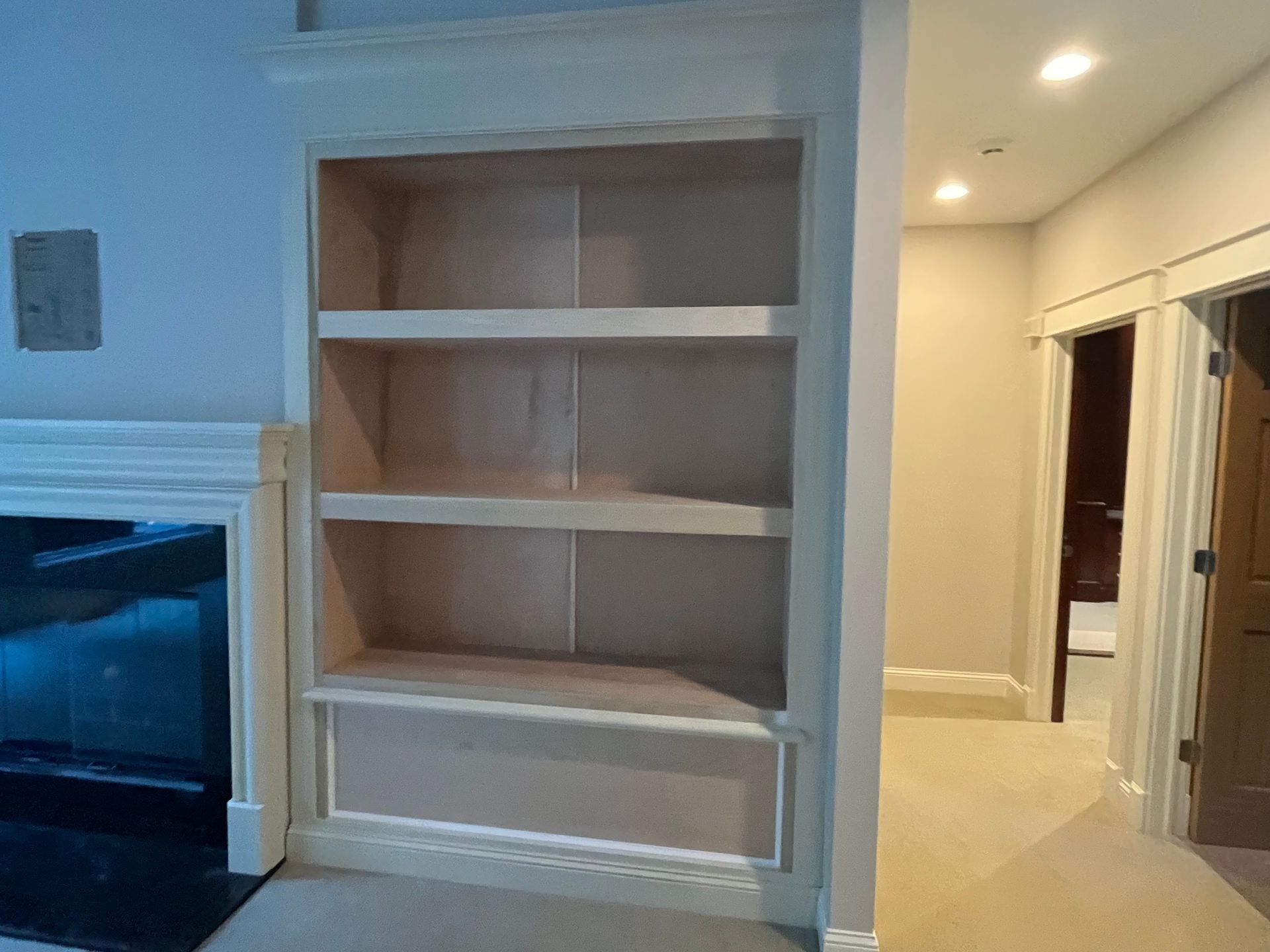 Built-in wooden bookshelf with three shelves and a recessed bottom, next to a fireplace, and a hallway.