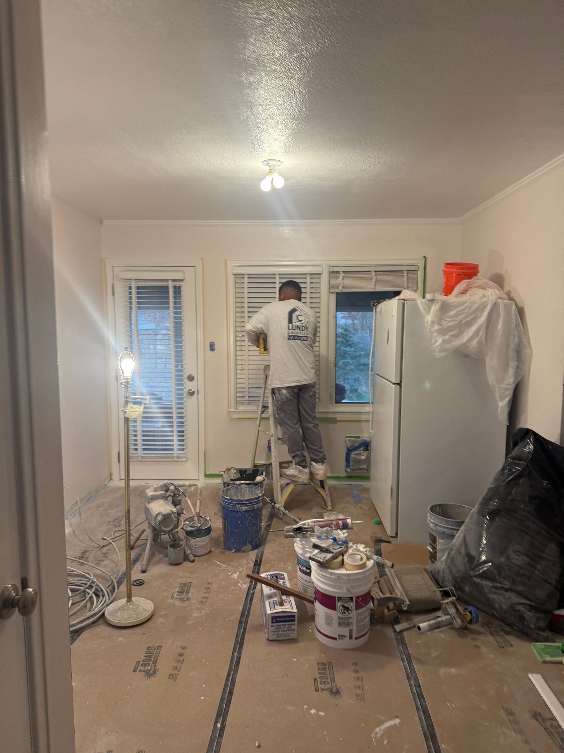 Person on ladder painting window trim in a room with debris on the floor and covered appliances.