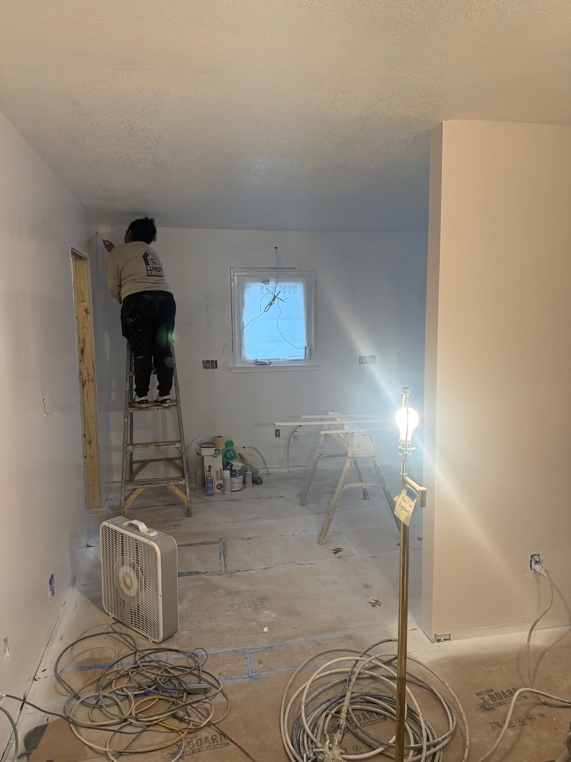Person on ladder painting a room during renovation. White walls, messy floor, tools, and wires visible.