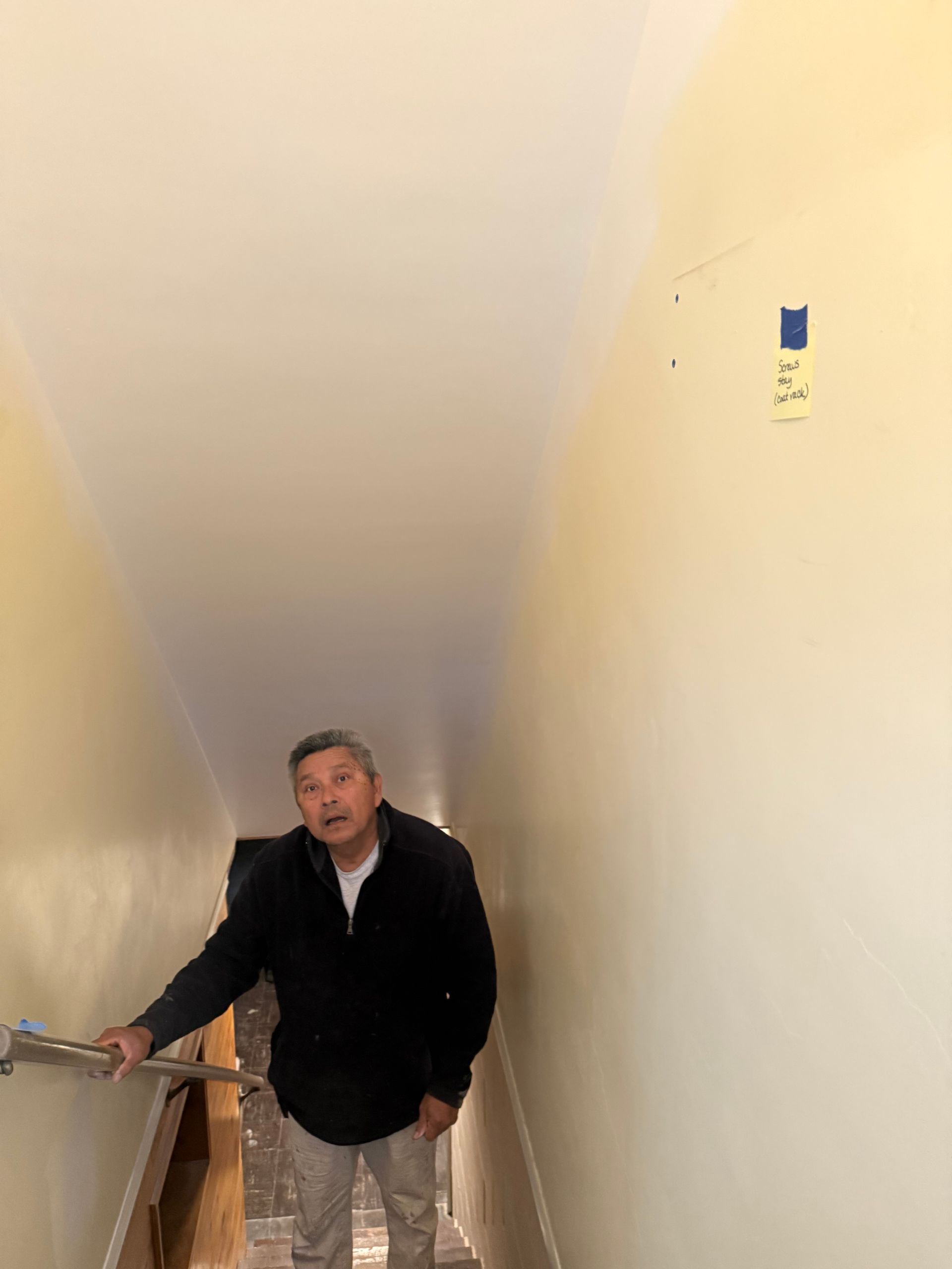 Man stands at the bottom of stairs, pointing towards the top; yellow walls, white ceiling, handrail.