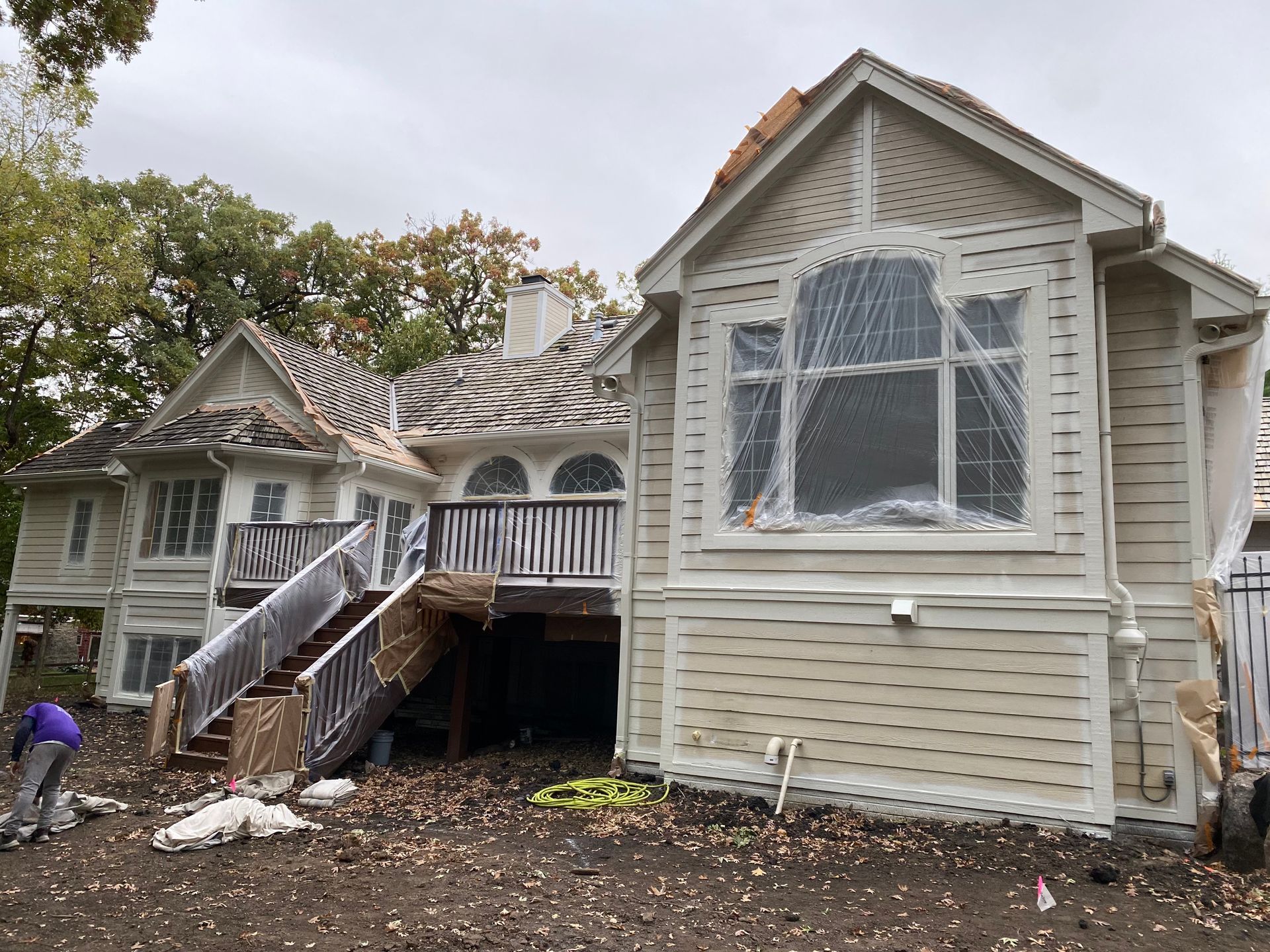 House under renovation, covered windows. Deck with stairs, person working on the ground. Overcast day.