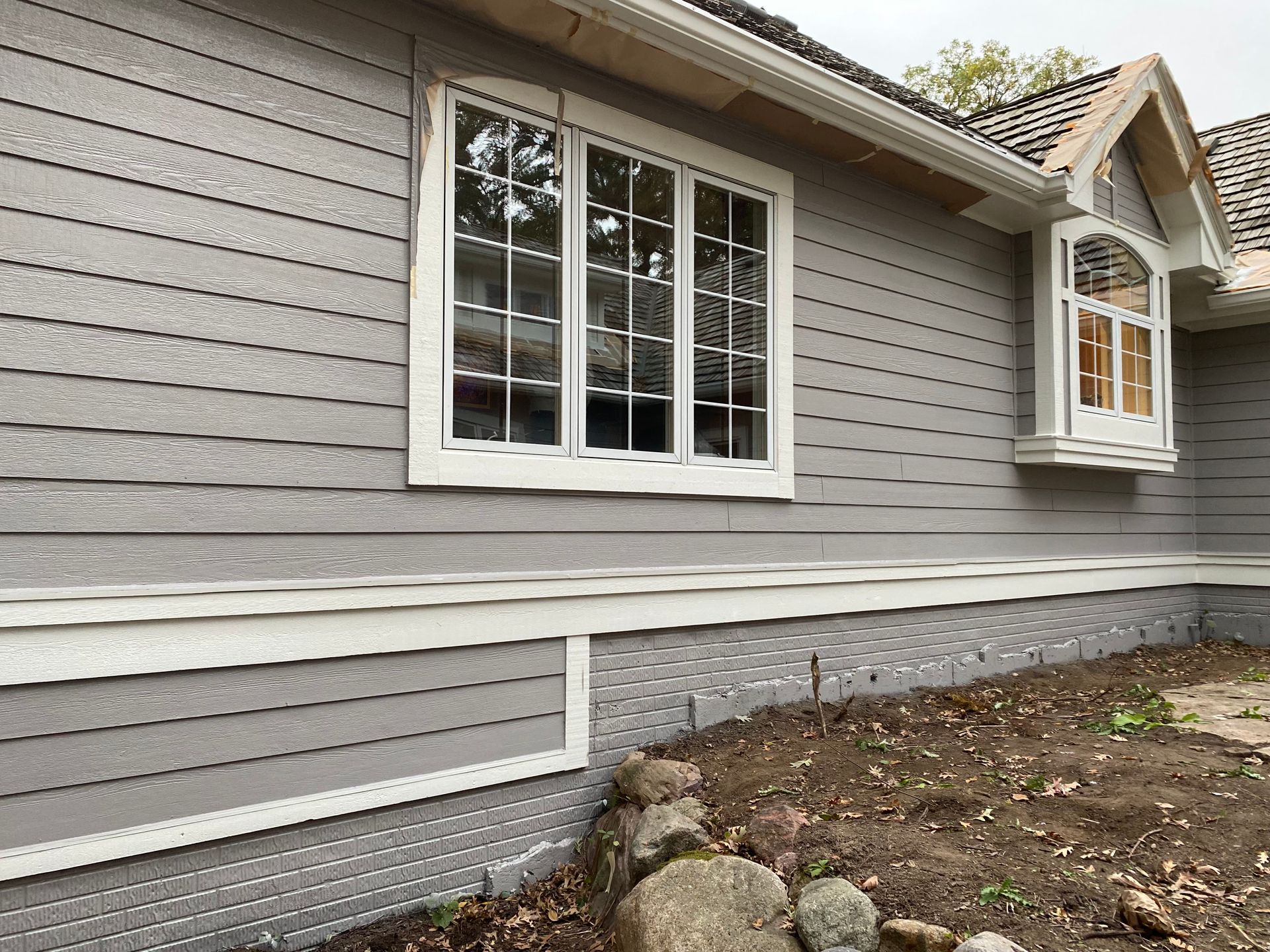 Gray siding on a house with white trim around windows. Dirt and rocks in front.