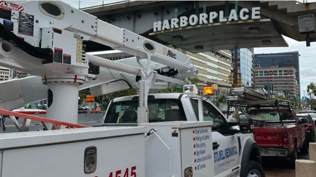 White utility truck with lift arm in front of Harborplace sign. Other trucks and buildings are visible.