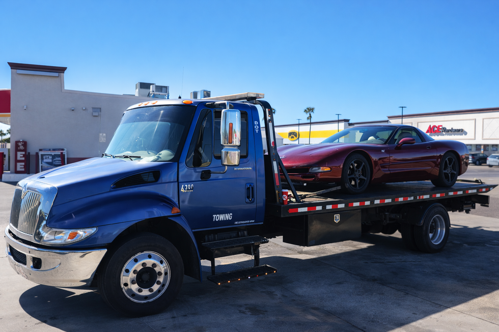 A blue flatbed tow truck transporting a maroon sports car in a parking lot on a sunny day.