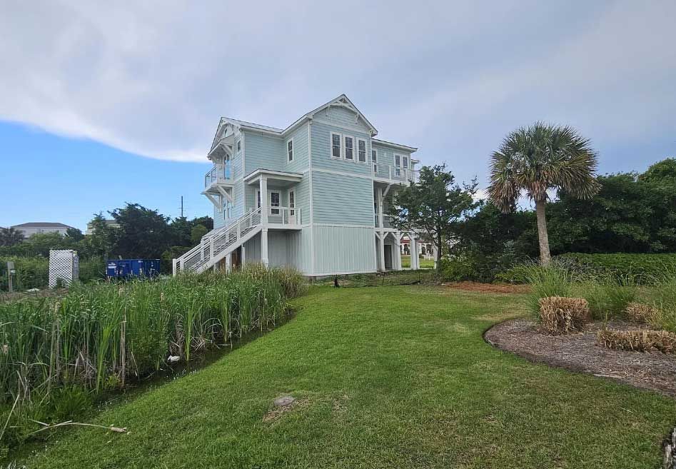 A light blue coastal house on stilts sits in a grassy yard near green vegetation and a palm tree under a cloudy sky.