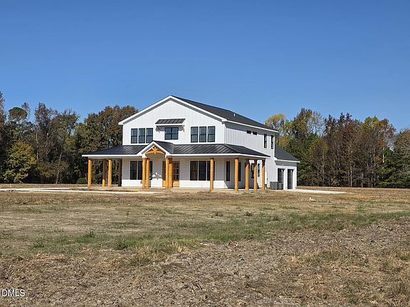 A two-story white farmhouse with a dark metal roof and covered porch stands on a large, grassy lot near a treeline.