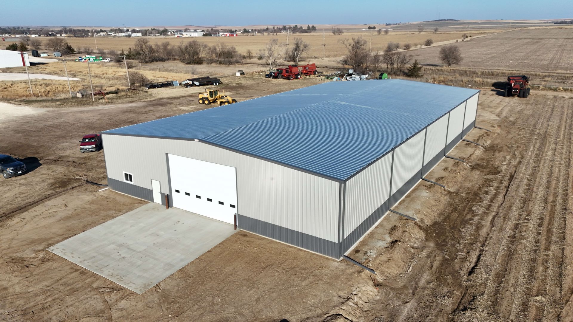 Large gray metal building with a white overhead door on a concrete pad, next to farmland.