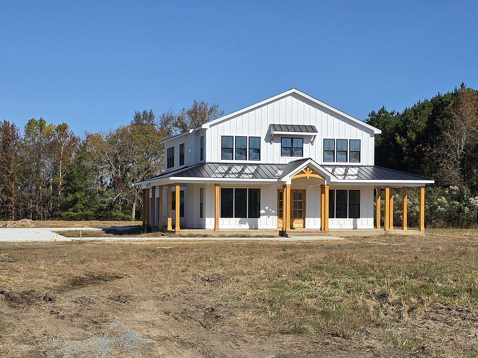 Two-story white house with a porch. Brown beams, gray roof, surrounded by trees and a dirt field under a blue sky.