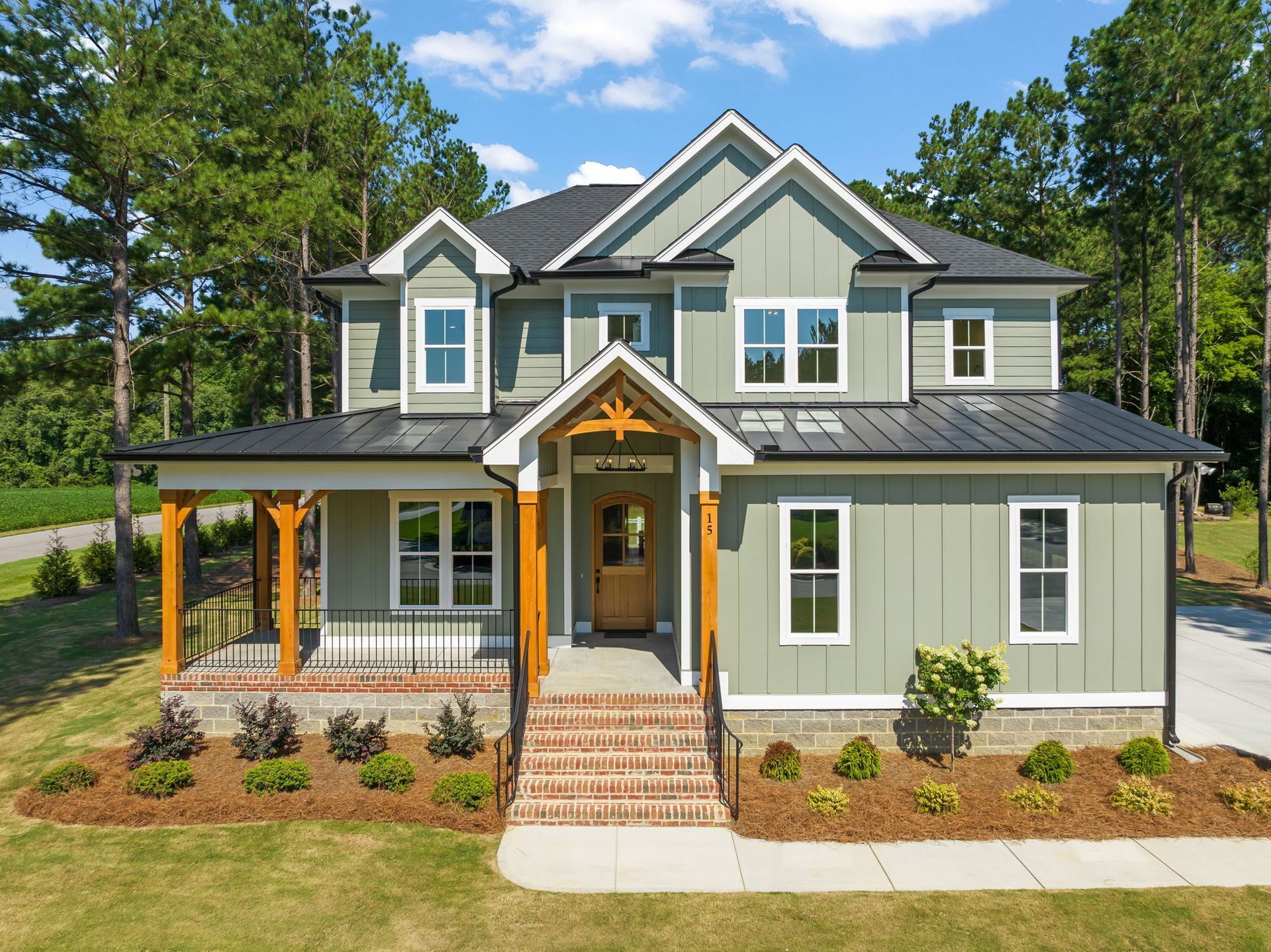 Two-story green house with black roof, wood porch, and brick steps, surrounded by trees and grass.