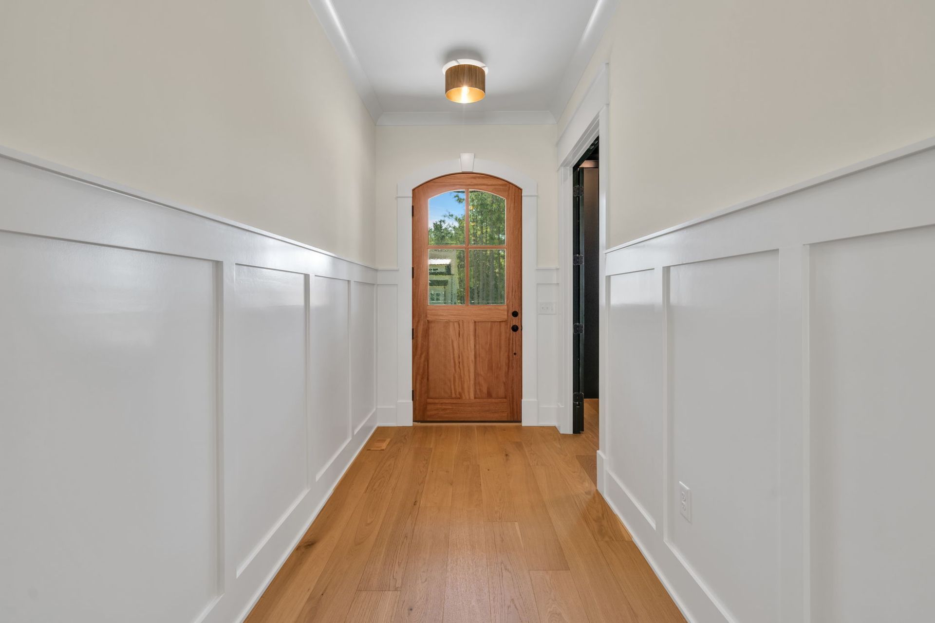 Hallway with white paneling, wooden floor, and a wood door with a window at the end.