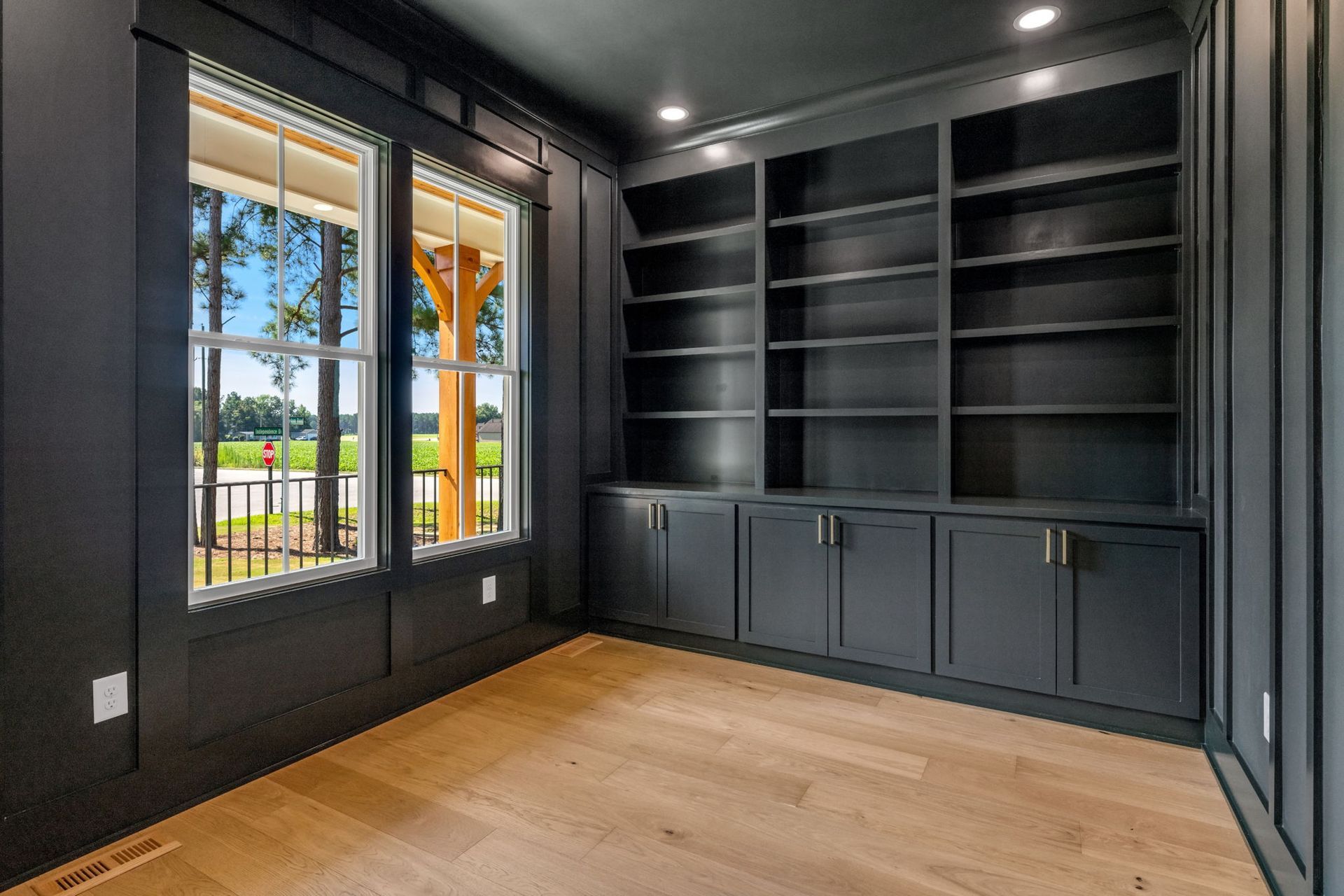 Dark-painted home office with built-in bookshelves and light wood floors. Windows overlook a yard with a view of trees.