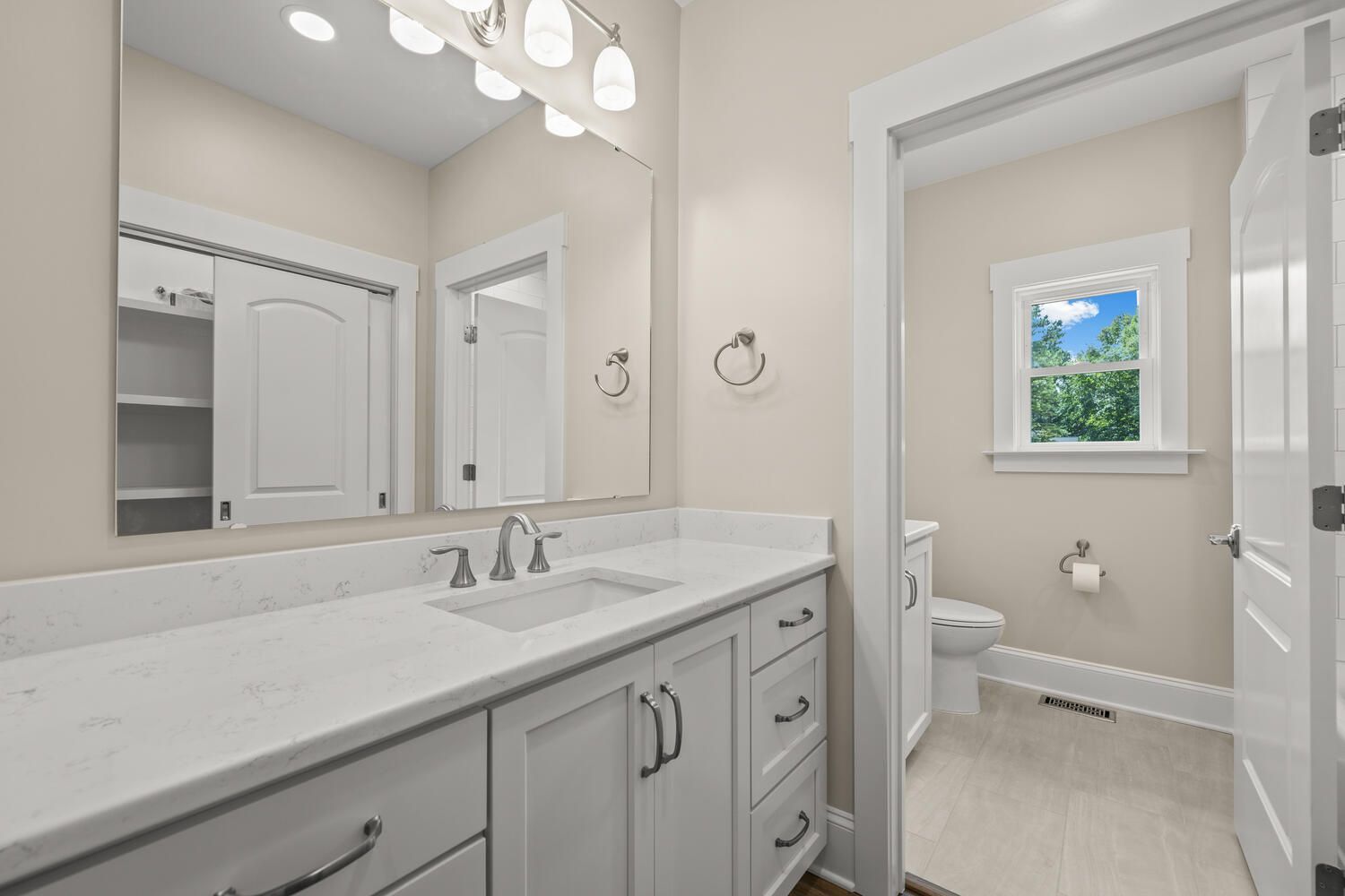 A bathroom with a sink, mirror, toilet, and window.