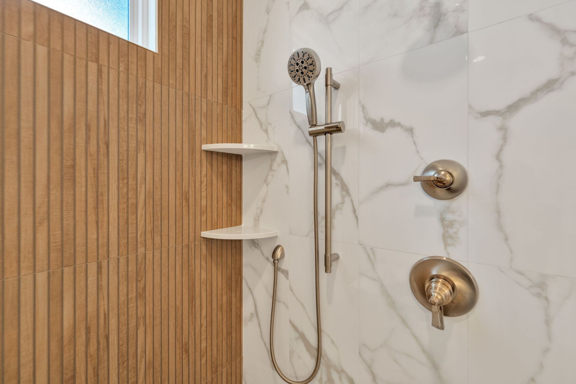 Shower with white marble walls, handheld showerhead, shelves, and wood-paneled wall.