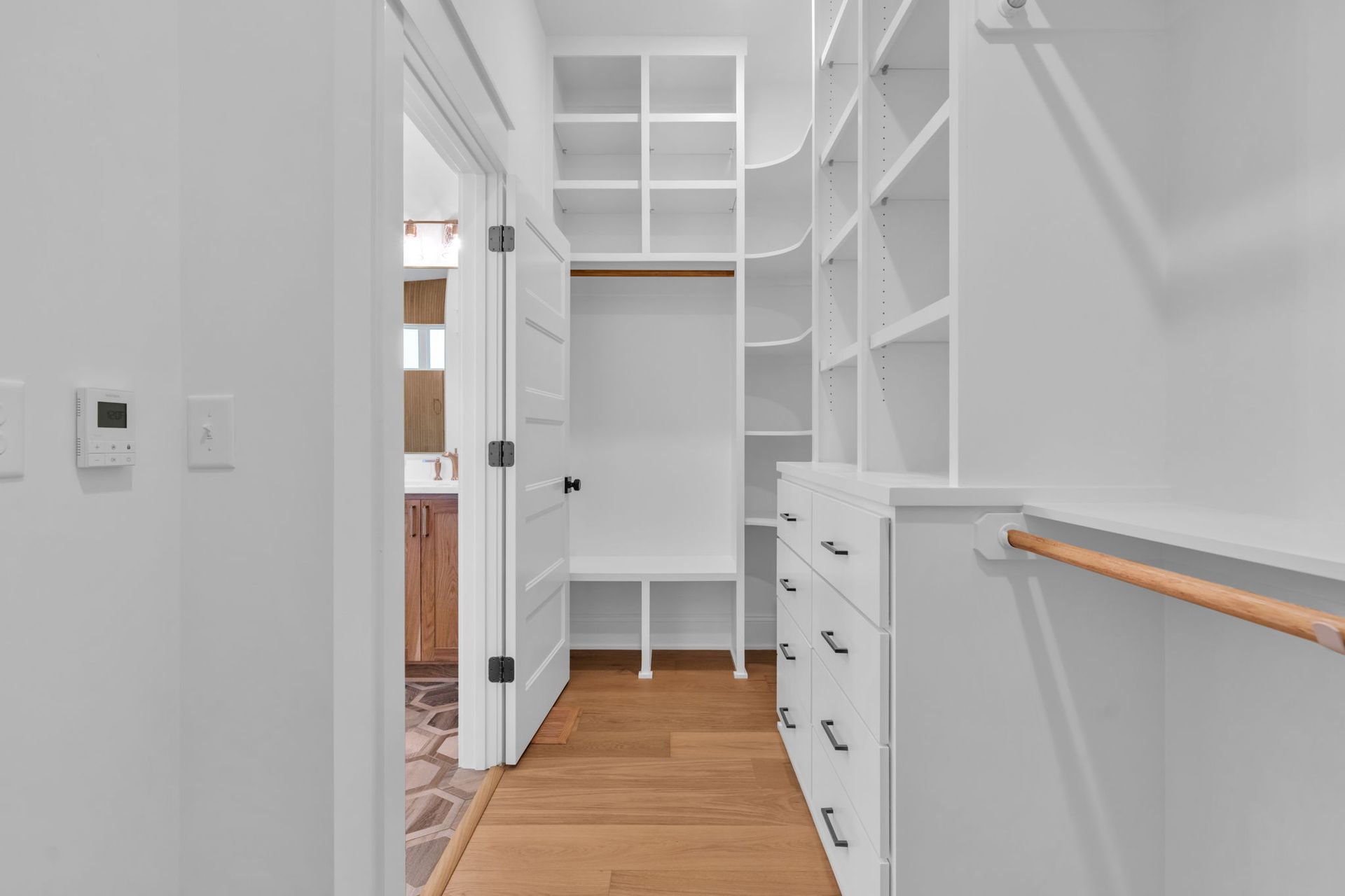 A bright white walk-in closet with wooden floors. Open door leads to a bathroom, shelves, and drawers line the walls.