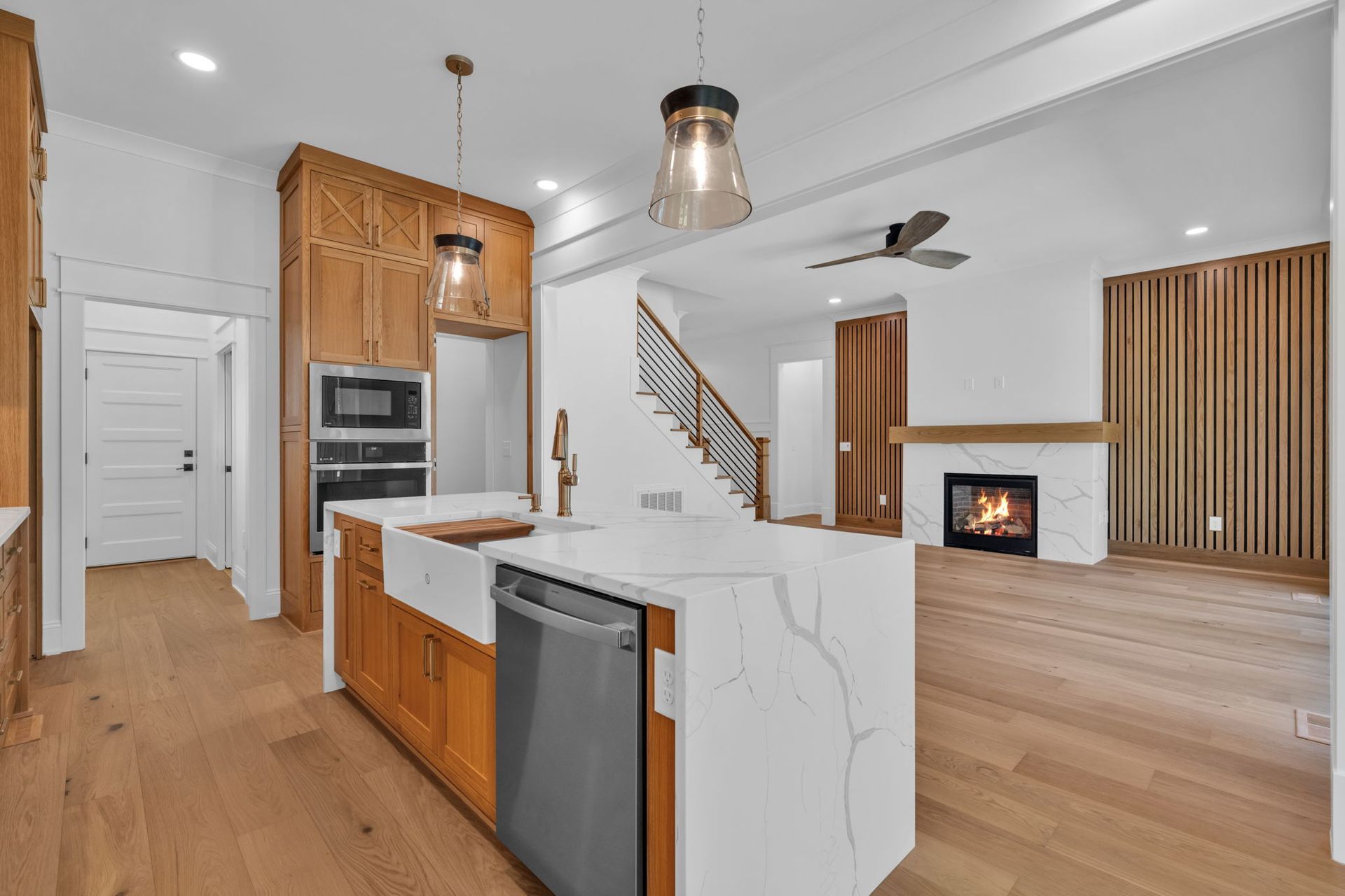 A modern kitchen with wooden cabinets and a white quartz island. A staircase and fireplace are visible in the background.