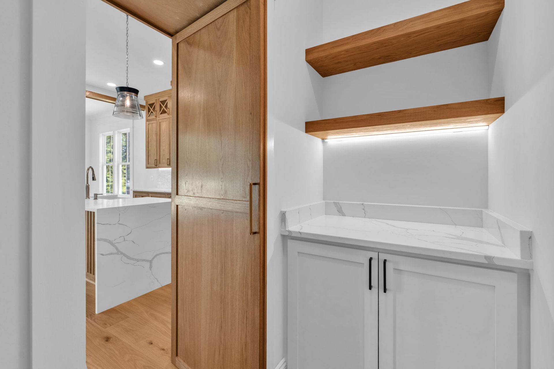 A bright kitchen pantry with white cabinets, shelves, and a wooden door; a kitchen island is visible.