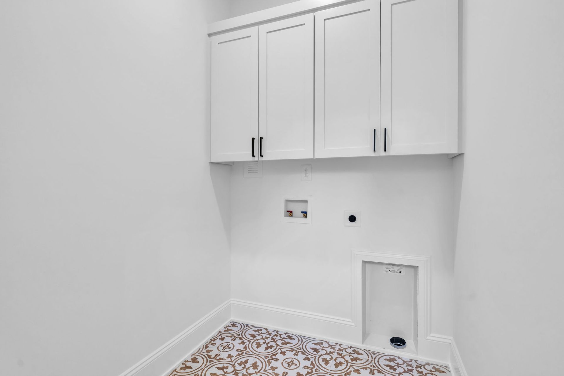 A white laundry room with upper cabinets, water hookups, and decorative floor tiles.