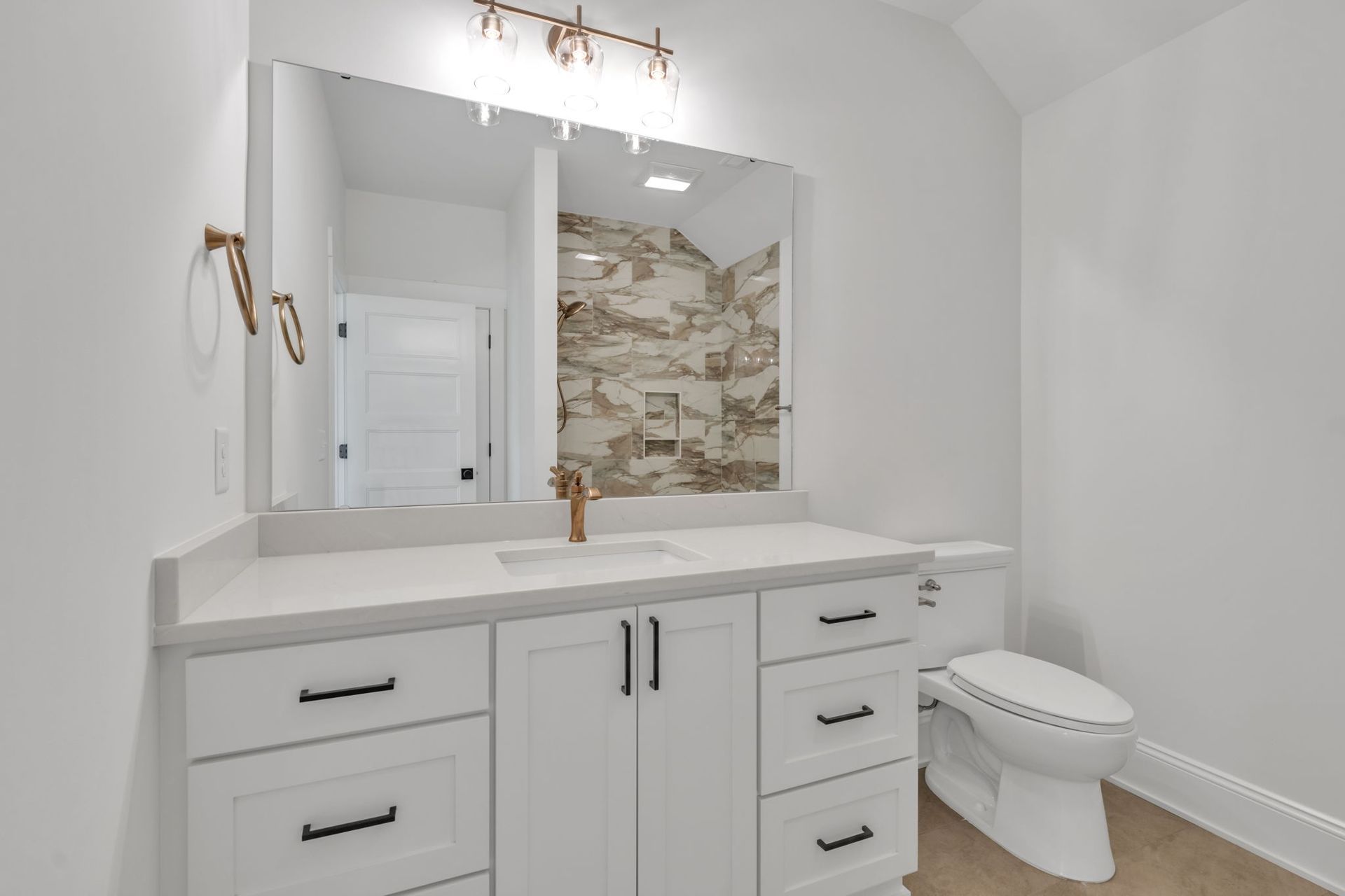 White bathroom with vanity, toilet, and mirror reflecting a shower. Gold fixtures and black hardware.