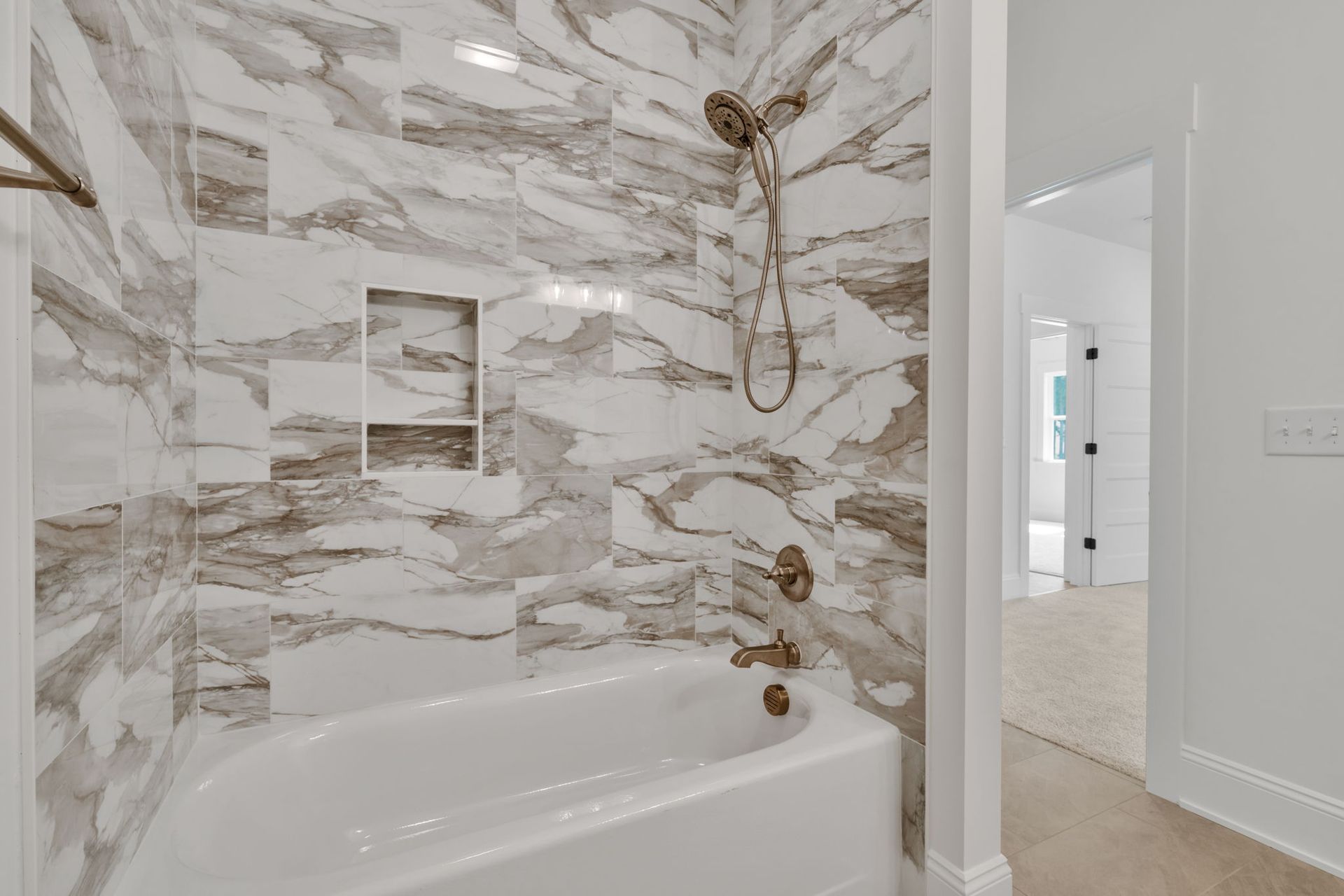 Bathroom with marble-patterned tiles, a built-in tub, and a showerhead. A doorway leads to a bedroom with a carpet.