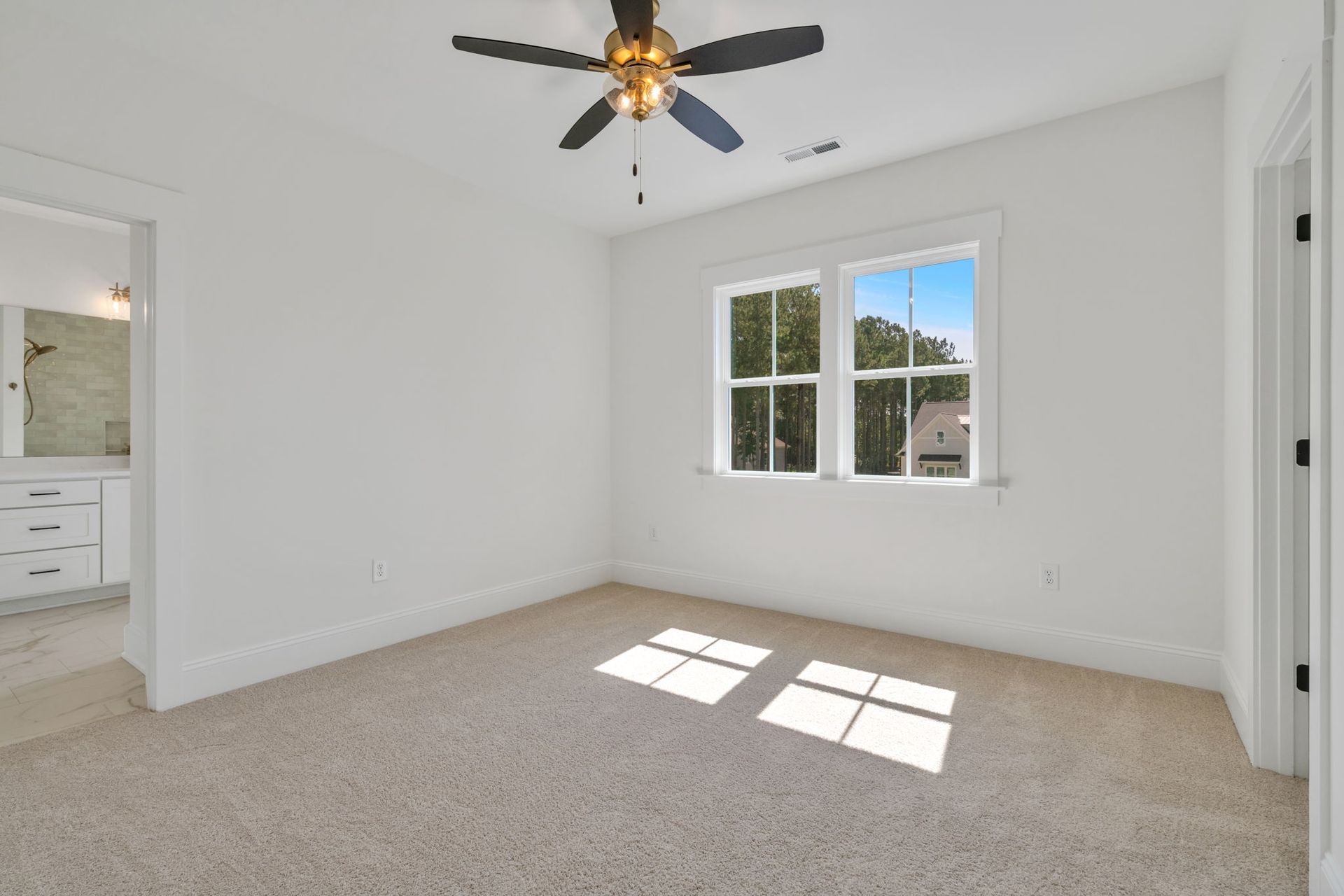 Empty bedroom with beige carpet, white walls, window, and ceiling fan. A bathroom is seen in the background.
