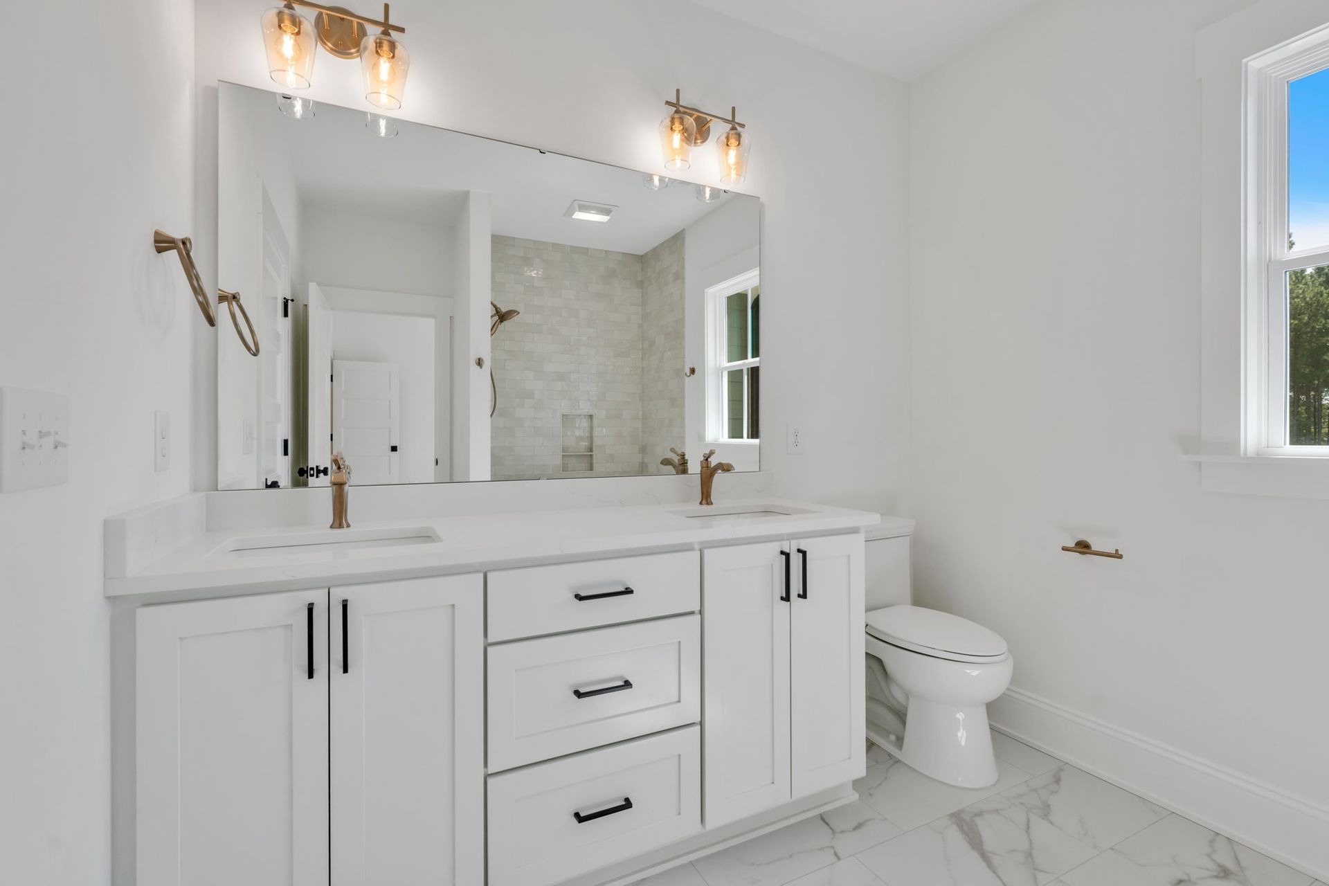 A bright white bathroom with a double vanity, large mirror, and toilet.  Gold fixtures and black drawer pulls add contrast.