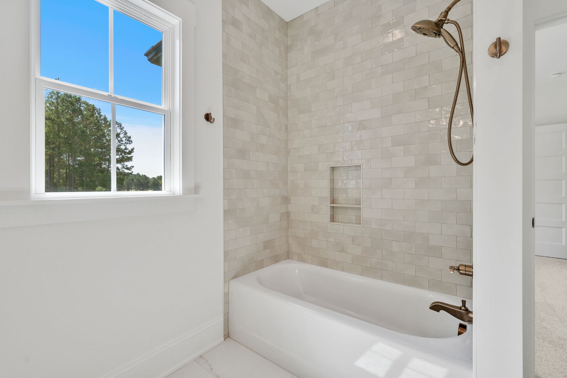 Bright bathroom with a white tub and tiled shower. A window offers a view of trees and sky.