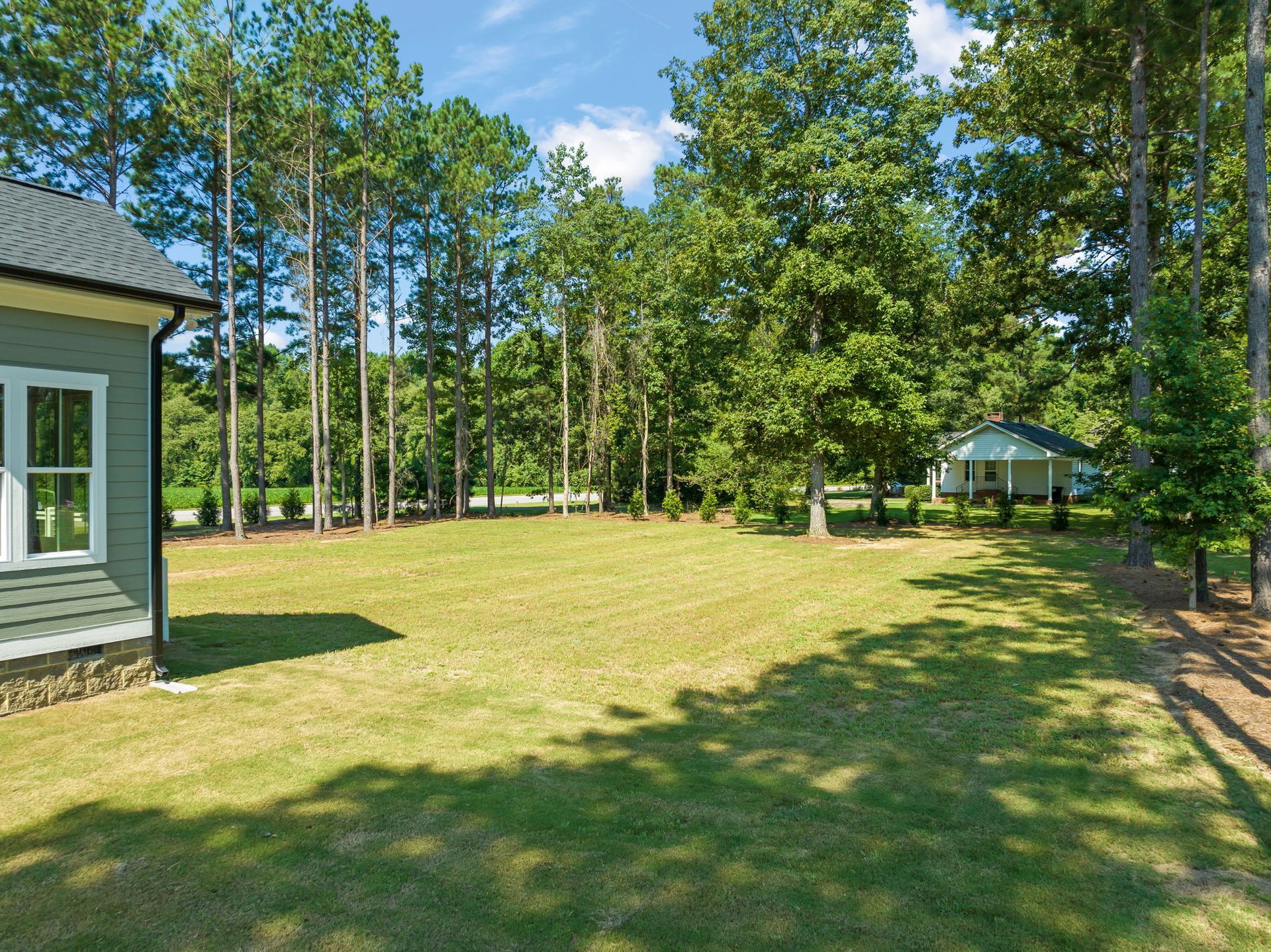 A grassy yard with trees in the background, with a house on the left and another in the distance on the right.