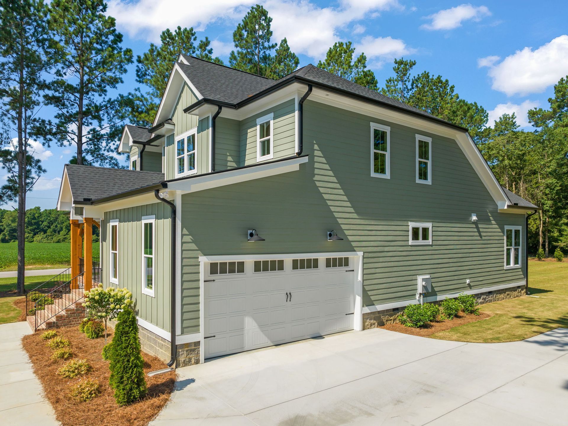Two-story, green house with a white garage door and a concrete driveway, surrounded by trees and a blue sky.