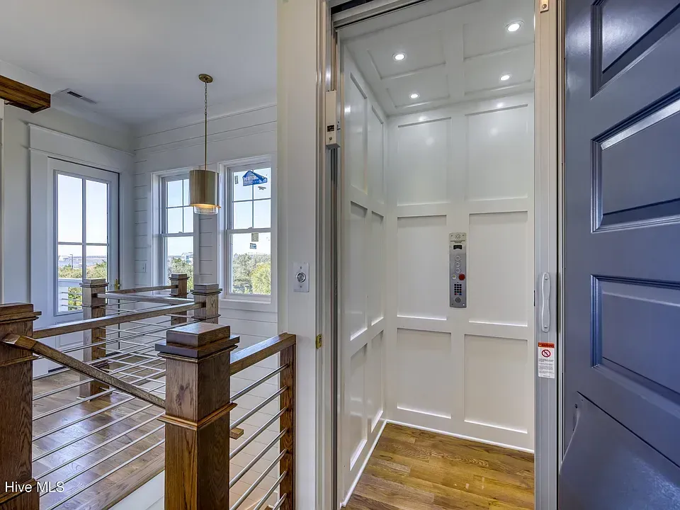 Elevator in a home's interior, white paneling, open door, wood flooring, staircase in view.