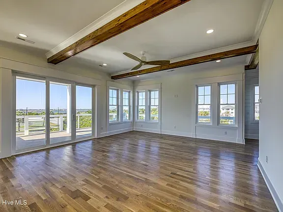 Empty room with wood floors, large windows, ceiling beams, and a ceiling fan.