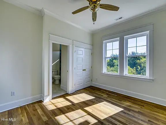 Bedroom with wood floors, two windows, a door to a small bathroom, and a closet. Light-colored walls and ceiling fan.