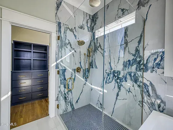 Bathroom with marble shower and a view into a walk-in closet with dark blue drawers and shelves.