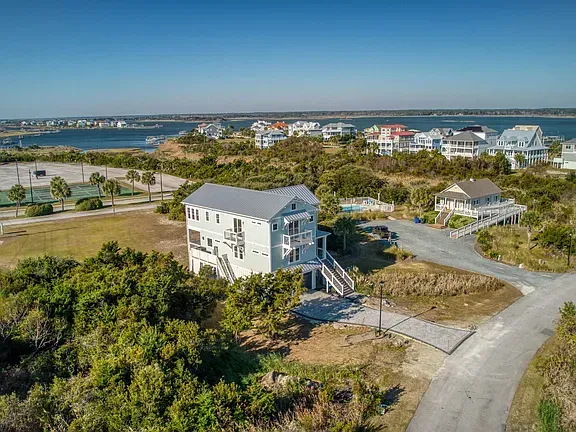 Coastal home with a light blue exterior and gray roof, surrounded by brush, with water and other houses visible.