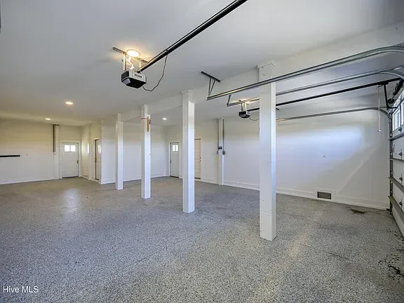 Empty garage with speckled floor, white walls, and support beams. Garage door is open.
