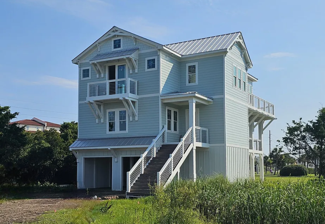Two-story blue beach house with white trim and stairs, on a grassy lot under a clear sky.