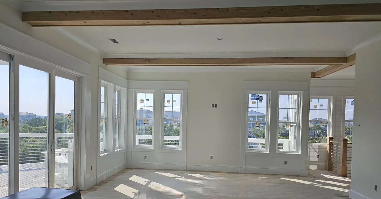 Bright room under construction with white walls, wooden beams, and large windows.
